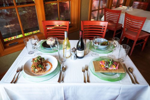 A beautifully set dining table with two plates of gourmet food, wine bottles, and elegant glassware.