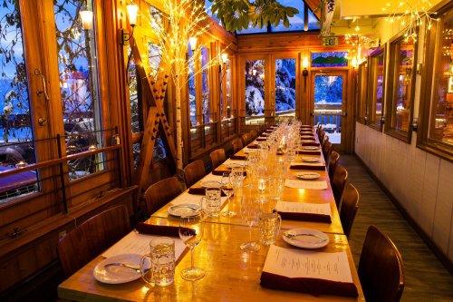 A warmly lit restaurant with a long wooden table set for dinner, surrounded by windows and festive lights.