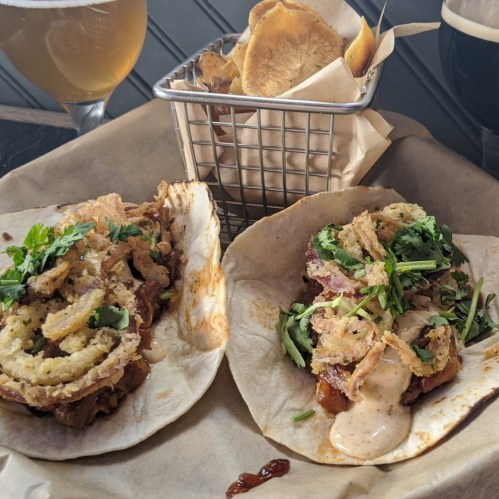 Two tacos topped with crispy onions and cilantro, served with a side of chips in a metal basket and two beers in the background.
