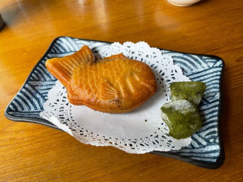 A fish-shaped pastry on a decorative plate, accompanied by two green sweets, on a wooden table.