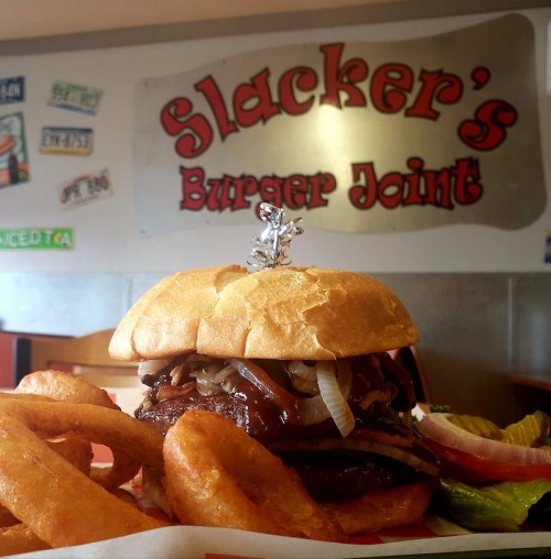 A delicious burger topped with onions and a small figure, served with onion rings, in front of a restaurant sign.
