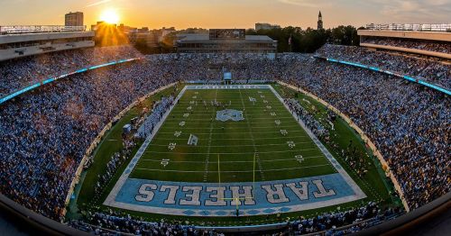 A panoramic view of a packed football stadium at sunset, with fans cheering and the field marked for the Tar Heels.