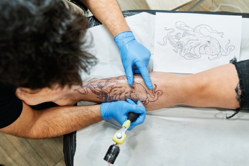 A tattoo artist works on an octopus design on a client's leg, with a reference sketch nearby.