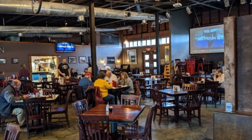 A spacious restaurant interior with wooden tables, patrons dining, and a large screen displaying a sports event.