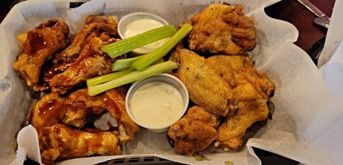 A basket of assorted chicken wings with two dipping sauces and celery sticks on a bed of parchment paper.