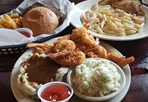 A plate of fried shrimp with mashed potatoes and coleslaw, alongside a burger, fries, and a pasta dish.