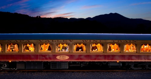 A vintage train car illuminated at dusk, with elegant dining setups visible through its windows against a mountain backdrop.