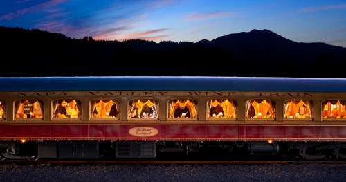 A vintage train car illuminated at dusk, with elegant dining setups and scenic mountains in the background.