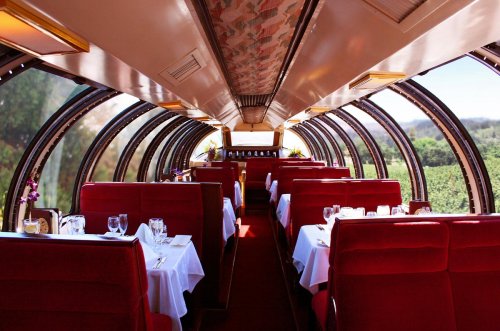 Interior of a dining train car with red seating, white tablecloths, and large windows showcasing a scenic view.
