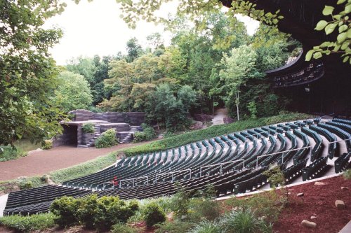An outdoor amphitheater surrounded by trees, featuring tiered seating and a stage area in the background.