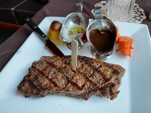 A grilled steak on a white plate, accompanied by sauces and a roasted potato.