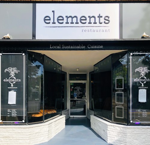 Exterior view of "elements" restaurant, featuring large glass windows and a sign promoting local sustainable cuisine.