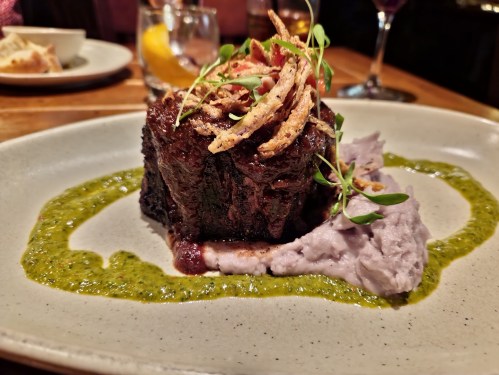 A plated dish featuring a piece of meat topped with crispy onions, served with purple mashed potatoes and green sauce.