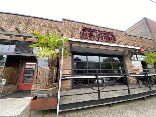 Exterior of Asado restaurant featuring a brick facade, large windows, and a palm tree in a planter.