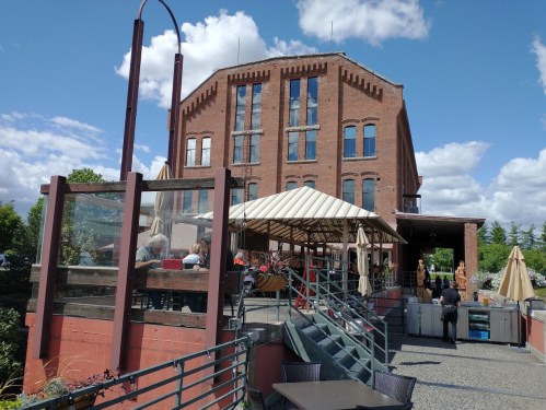 A brick building with large windows, featuring an outdoor dining area with umbrellas and a staircase leading up.