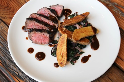 Sliced steak with a rich sauce, served with roasted potatoes and sautéed greens on a white plate.