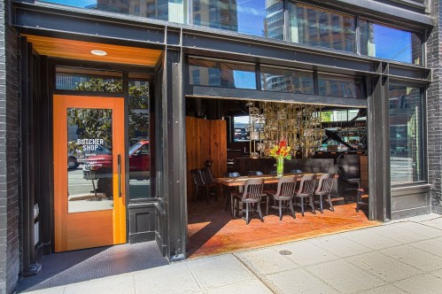 Exterior of a butcher shop with large windows, showcasing a dining area and a modern chandelier inside.