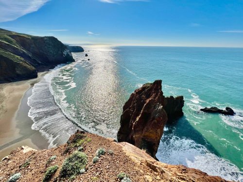 A scenic coastal view featuring cliffs, a sandy beach, and gentle waves under a clear blue sky.