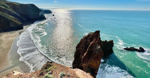 A scenic coastal view with cliffs, sandy beach, and sparkling blue water under a clear sky.