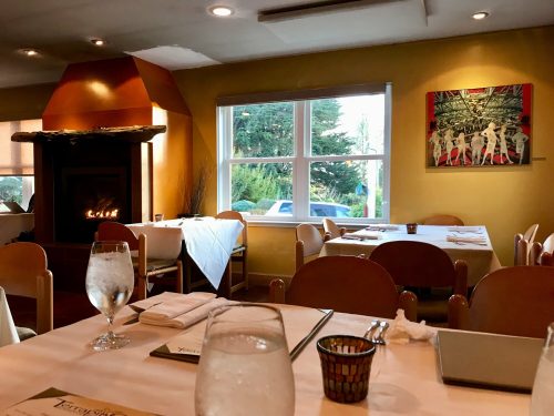 Cozy restaurant interior with tables set for dining, a fireplace, and a window showing greenery outside.