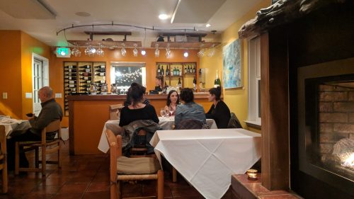 Cozy restaurant interior with a bar, tables set for dining, and a fireplace, featuring patrons enjoying their meals.