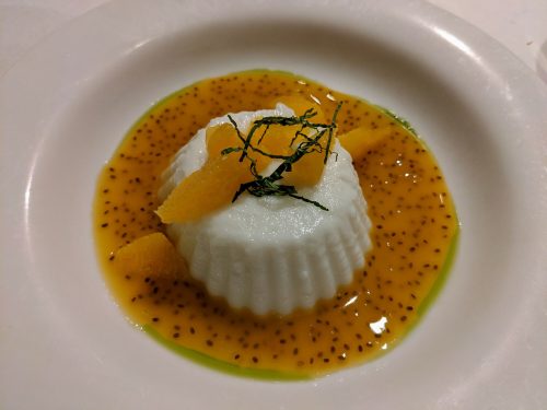 A creamy dessert topped with mango sauce, mint leaves, and pieces of fruit, served on a white plate.