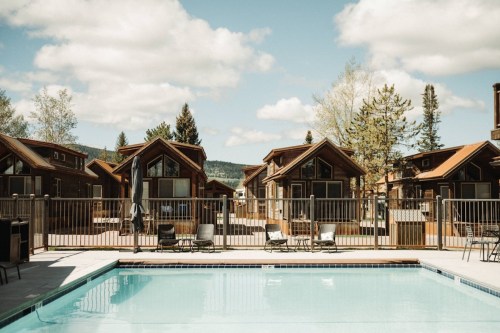 A serene pool area surrounded by wooden cabins and trees under a blue sky with fluffy clouds.