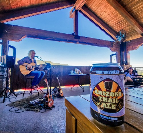A musician performs with an acoustic guitar on a patio, with a can of Arizona Trail Ale in the foreground.