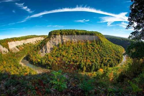 A panoramic view of a river winding through colorful autumn foliage and steep cliffs under a blue sky with wispy clouds.