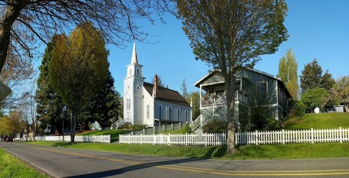 A scenic view of a white church and a house beside a tree-lined road, with a white picket fence in the foreground.