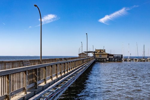 A wooden pier extends over calm water, leading to a building with a yellow roof and sailboats in the background.