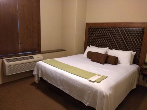 Cozy hotel room featuring a queen bed with white linens, a green throw, and a window with wooden blinds.