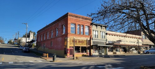 Historic brick buildings line a street under a clear blue sky, with parked cars and trees in the foreground.