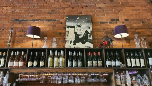 A wooden shelf displaying various wine bottles, with a black-and-white portrait of a woman above and lamps on either side.