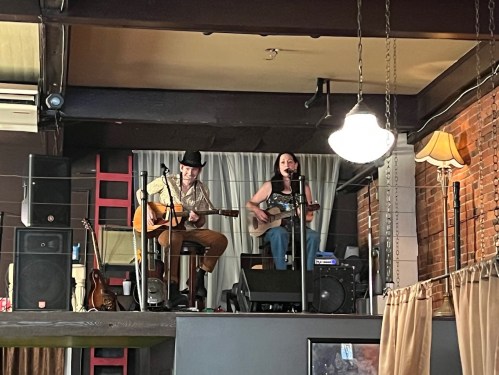 A man and woman perform music on a stage in a cozy venue, surrounded by instruments and warm lighting.