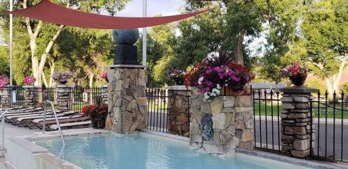 A serene pool area with stone features, colorful flowers, and shaded lounge chairs surrounded by greenery.