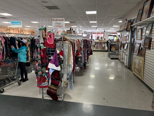 A spacious thrift store aisle filled with clothing racks and holiday decorations, with a shopper browsing nearby.