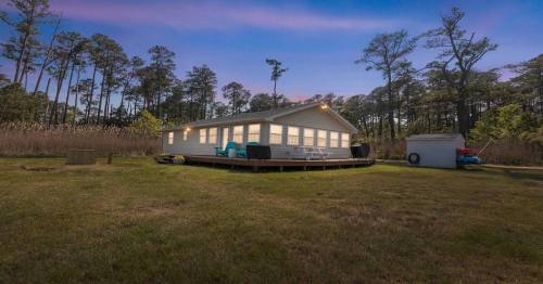 A cozy white house with large windows, surrounded by trees and tall grass, set against a twilight sky.