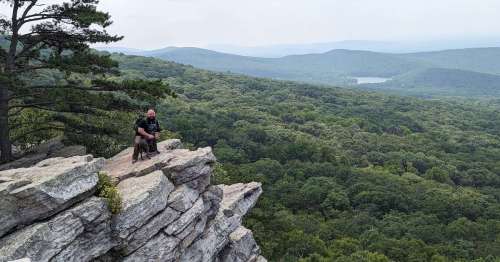 A person and a dog sit on a rocky ledge, overlooking a lush green valley and distant mountains under a cloudy sky.