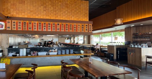 Interior of a modern restaurant with wooden decor, a sushi bar, and a menu displayed on the wall.