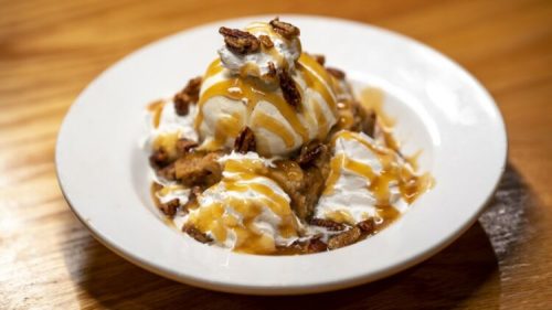 A dessert bowl with vanilla ice cream, whipped cream, caramel sauce, and crushed pecans on top.