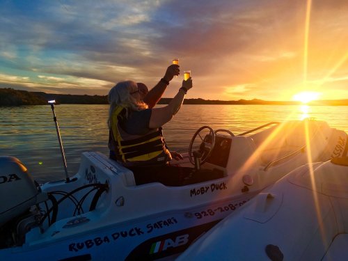 A person in a boat raises two glasses in celebration against a sunset over calm waters.