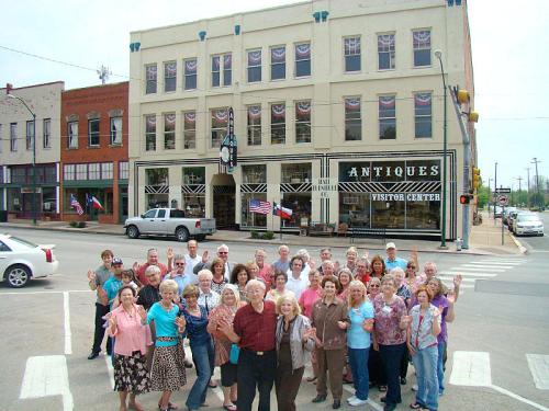 A large group of smiling people stands in front of an antique store, waving at the camera on a sunny day.