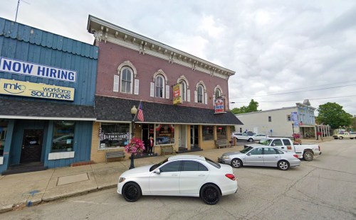 A street view of a two-story brick building with shops, a barber shop, and parked cars under a cloudy sky.