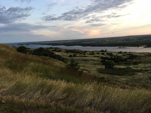A scenic view of rolling hills and a river at sunset, with a colorful sky and lush greenery.