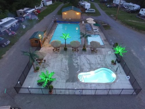 Aerial view of a pool area with two pools, lounge chairs, umbrellas, and illuminated palm trees at a campsite.