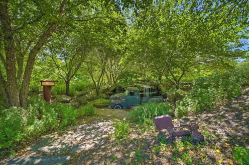 A vintage truck nestled among lush greenery and trees, with a small waterfall and rustic seating nearby.