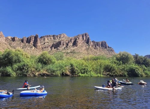 People paddle on inflatable rafts and paddleboards in a river surrounded by rocky cliffs and greenery under a clear blue sky.