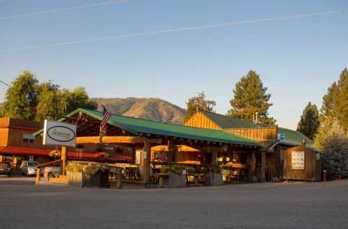 A rustic wooden building with a green roof, surrounded by trees and mountains, featuring outdoor seating and signage.