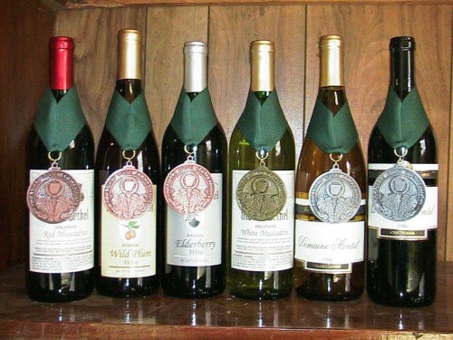 A row of six wine bottles with labels and medals hanging from their necks, displayed against a wooden background.